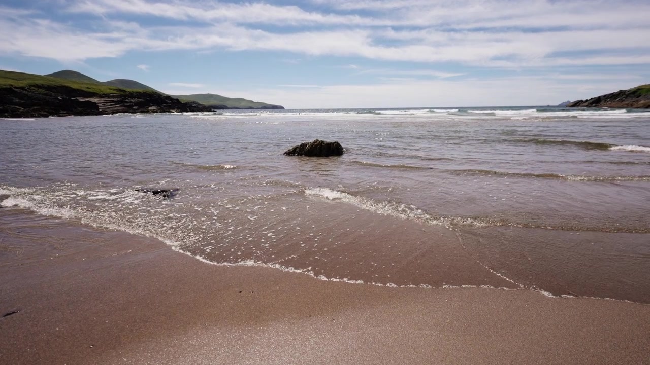 Dolci onde oceaniche si infrangono su una spiaggia sabbiosa con rocce sparse lungo un paesaggio costiero sotto un cielo parzialmente nuvoloso.