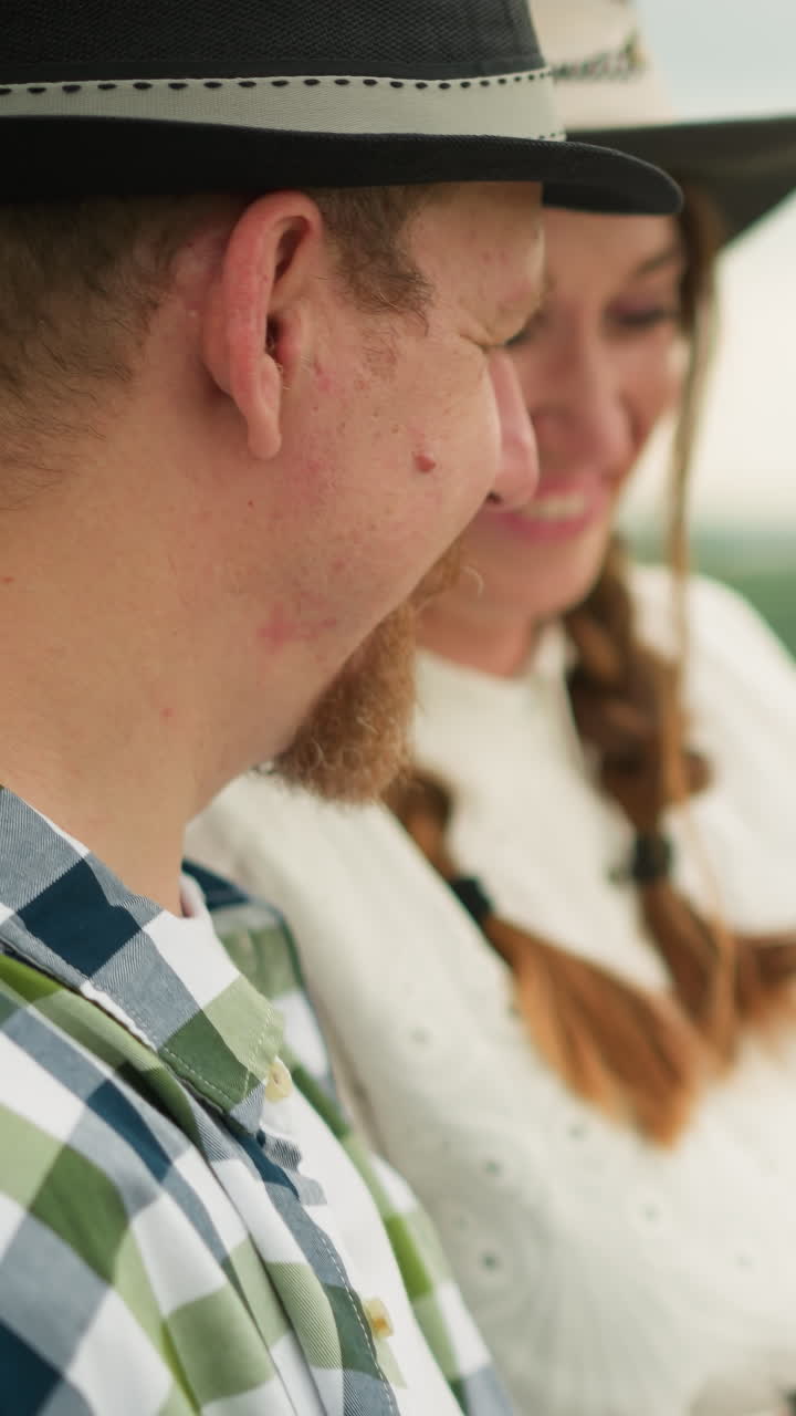primer plano de una pareja en un campo cubierto de hierba, con el hombre con un sombrero negro y una camisa a cuadros, y su pareja en un vestido blanco. ella sonríe cálidamente mientras le entrega un conjunto de pinceles