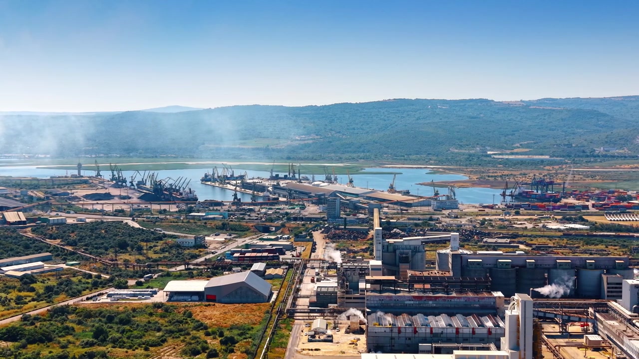 Luftperspektiv av havneterminal og omkringliggende industrianlegg. Lastkraner og skip ligger ved kaien i en bukt med grønne åser i bakgrunnen under en disig blå himmel.