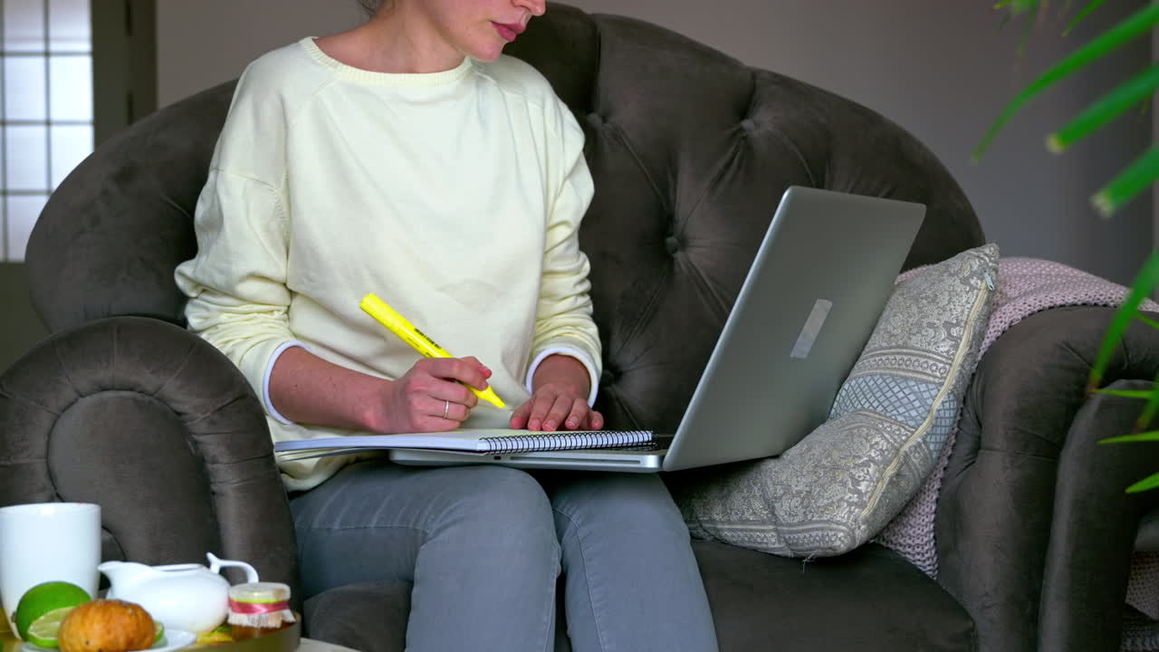Caucasian woman sitting on a chair, working on her laptop and making notes in notepad. Table with tea and fruits nearby