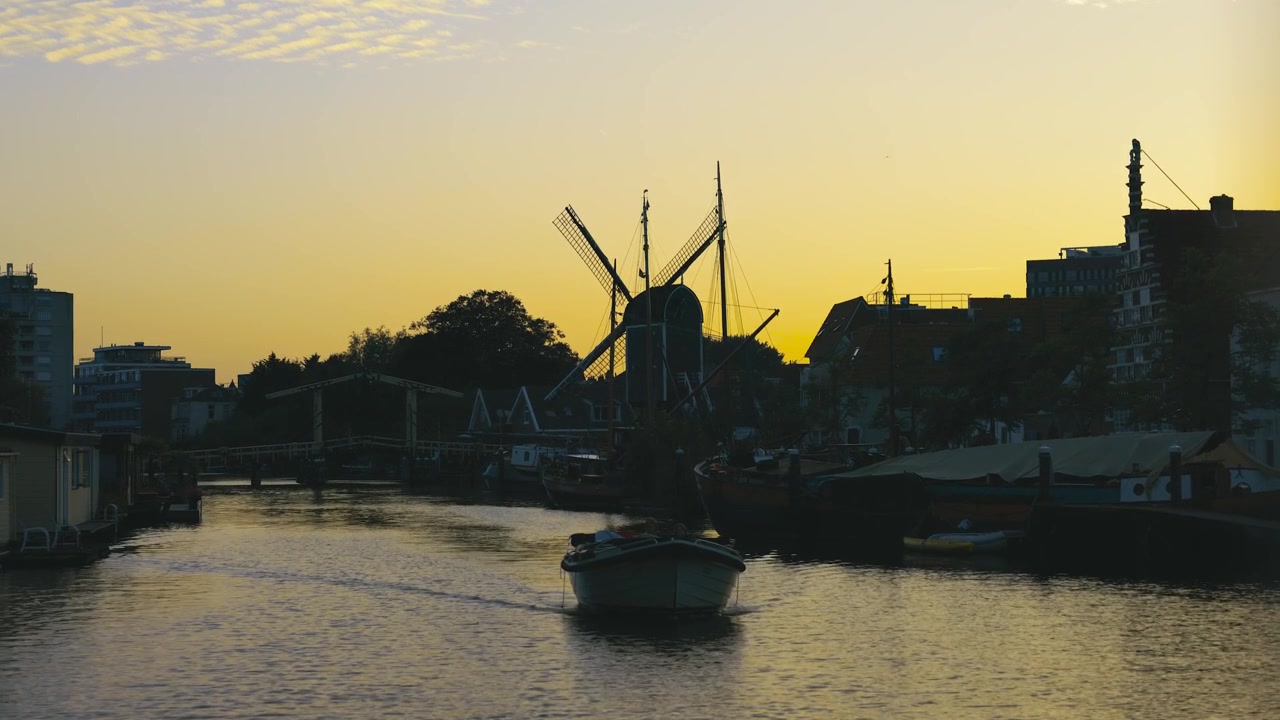 Silhouette di un mulino a vento tradizionale accanto a un canale calmo durante un tramonto dorato, mentre una piccola barca si muove sull'acqua creando un riflesso pacifico in un suggestivo paesaggio olandese.