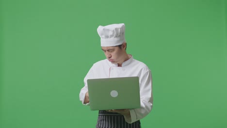 un chef asiático usando una computadora portátil y con dolor de cabeza mientras está de pie en el estudio de fondo de la pantalla verde