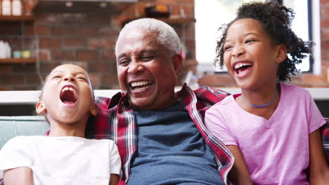portrait of grandfather sitting on sofa at home with granddaughters and laughing