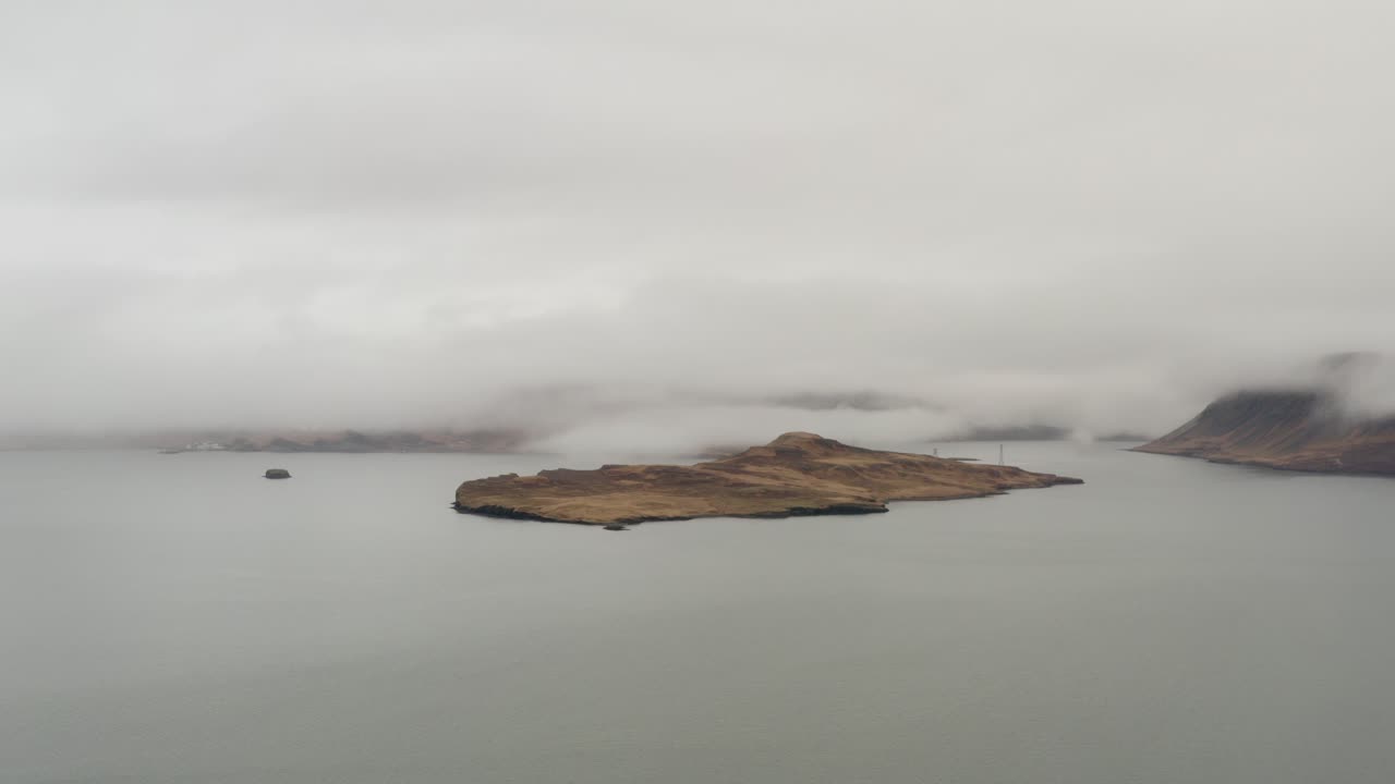 Vista aerea di un'isola avvolta dalla nebbia e circondata dalle acque calme di Hvalfjörður, in Islanda, con nuvole basse che aleggiano sopra la costa e una luce soffusa attraverso il cielo nuvoloso