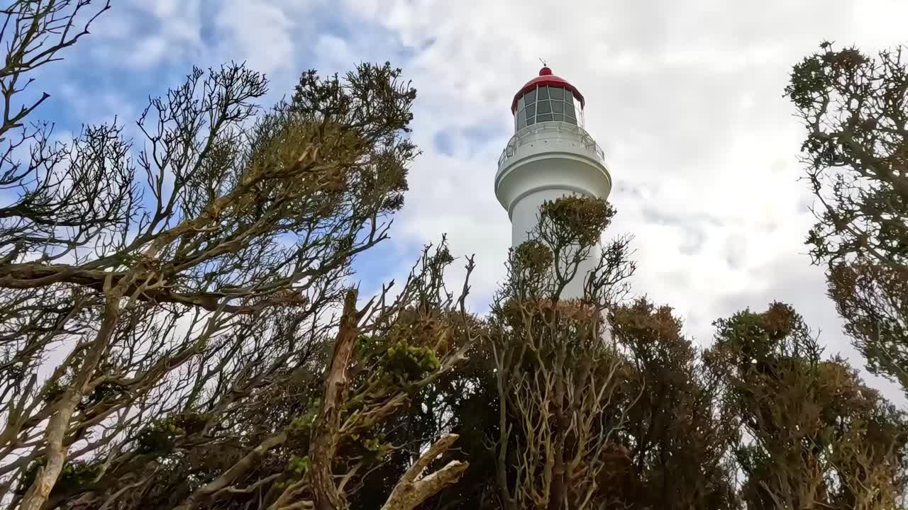 A lighthouse rises above windswept trees under a partly cloudy sky, showcasing a coastal landscape.