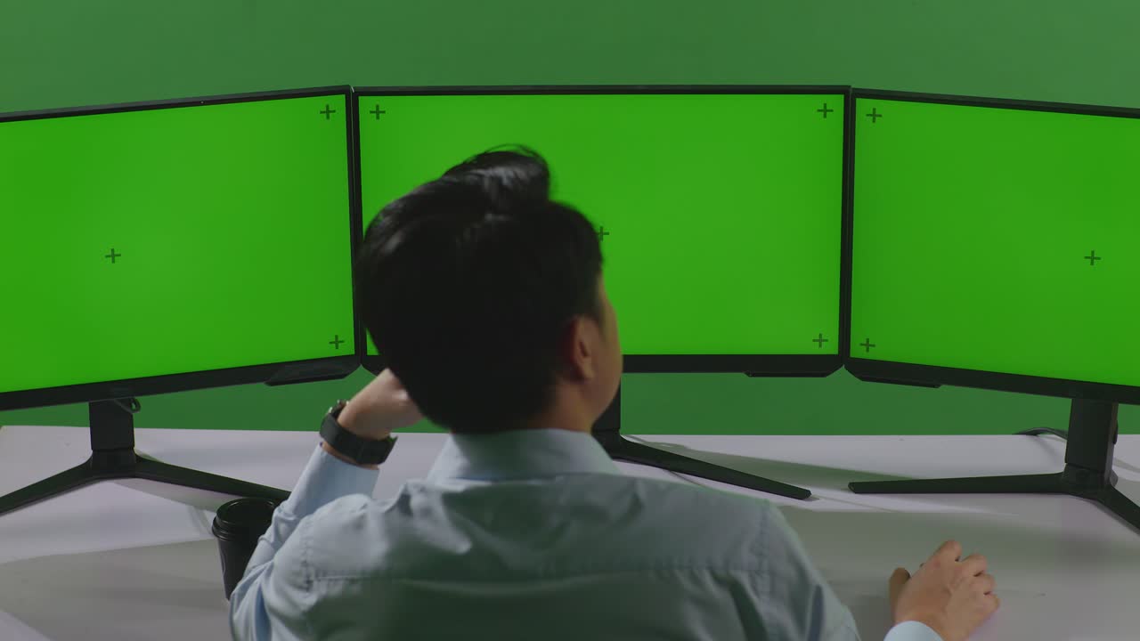 지루한 아시아인 남자가 녹색 스크린 스튜디오에서 여러 컴퓨터 모니터를 모방하여 작업하는 뒷면