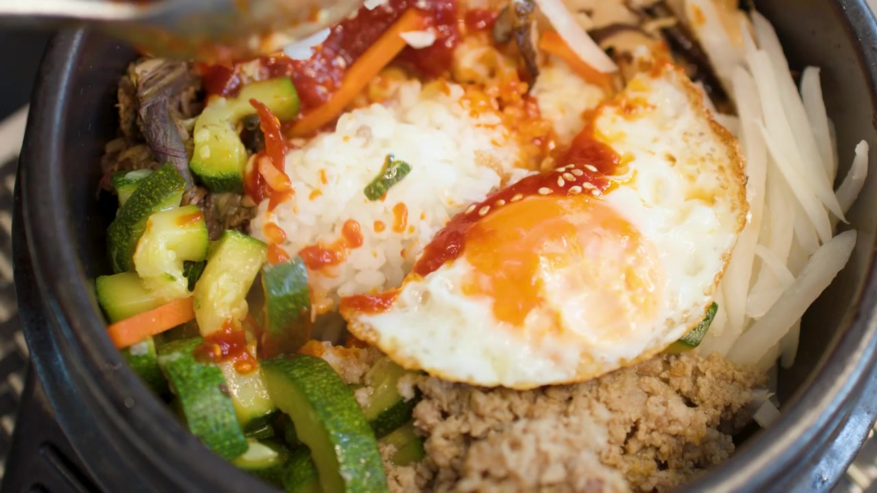 Sebuah sendok mengaduk nasi, sayuran, dan telur goreng dalam mangkuk batu dolsot panas