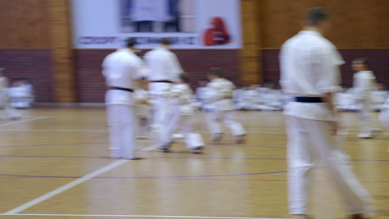 Little sportsmen split into pairs to practice karate fight. Coaches watch every pair of athletes. Other kids waiting for their turn sitting on the floor at backdrop.