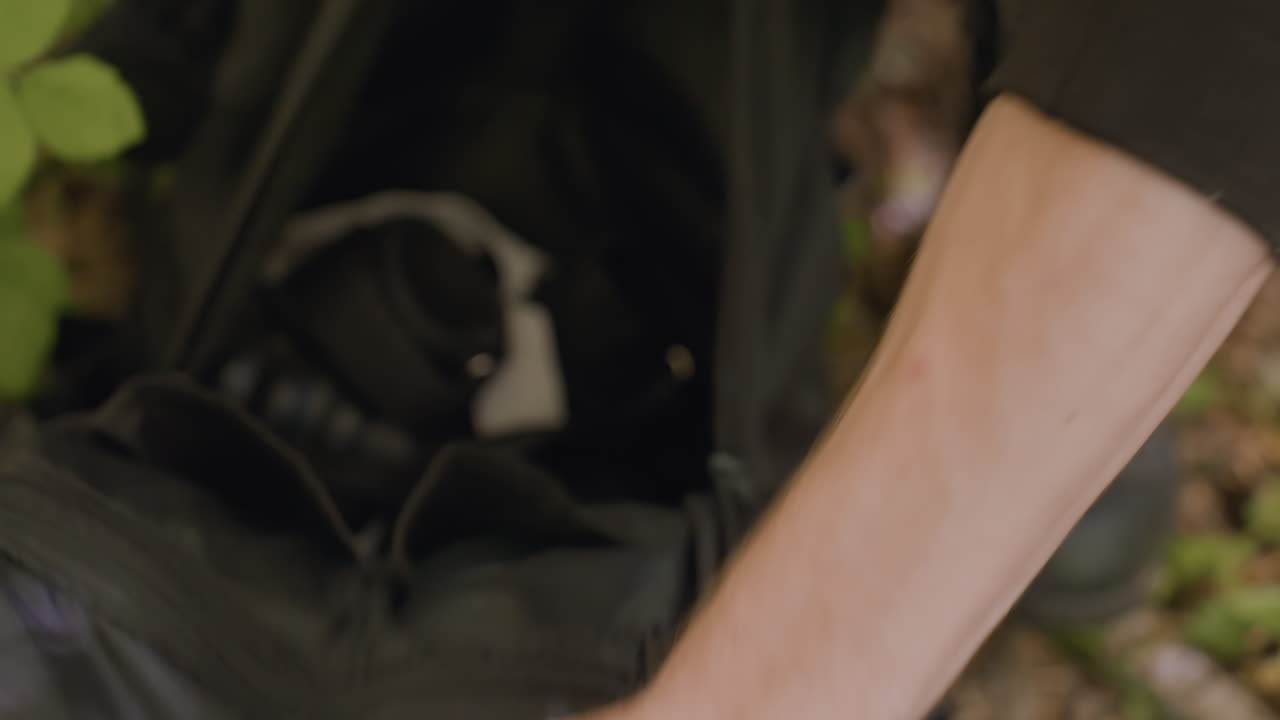 Young man unzips bag in forest, hands pulling zipper open as green leaves brush against bag, revealing blurry view of contents including gear, dirt-covered trousers, and natural surroundings
