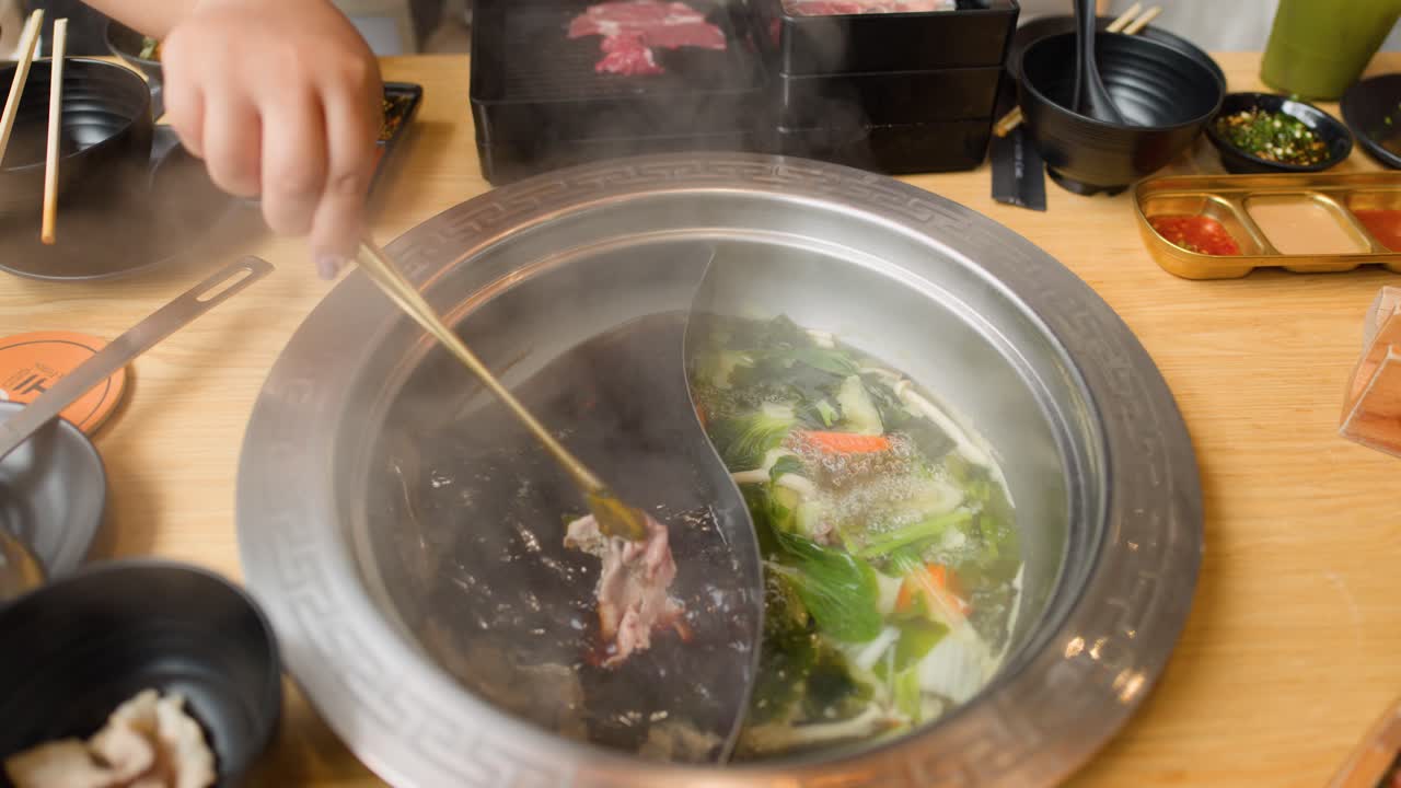 Hand cooks sliced beef in steaming dual broth hotpot, fresh vegetables, overhead view, natural lighting