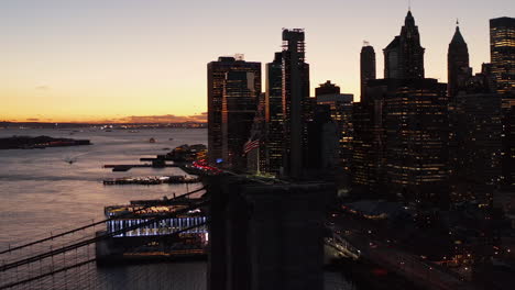 Orbit-shot-around-American-flag-on-top-of-suspension-tower-of-Brooklyn-bridge.-Panning-shot-of-city-at-dusk.-Skyscrapers-against-sunset-sky.-New-York-City,-USA