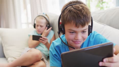 retrato de un hermano y una hermana caucásicos acostados en el sofá usando tableta y teléfono inteligente