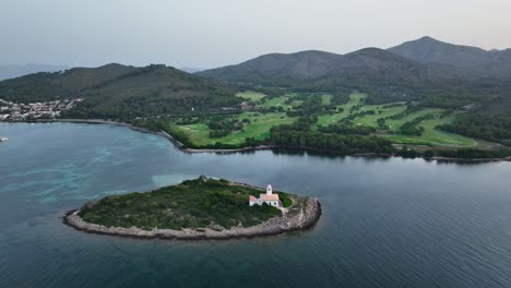 Drone-revealing-lighthouse-on-small-island-in-crystal-clear-water-next-to-beautiful-Alcanada-golf-course-on-Mallorca
