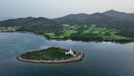 drone revealing lighthouse on small island in crystal clear water next to beautiful alcanada golf course on mallorca