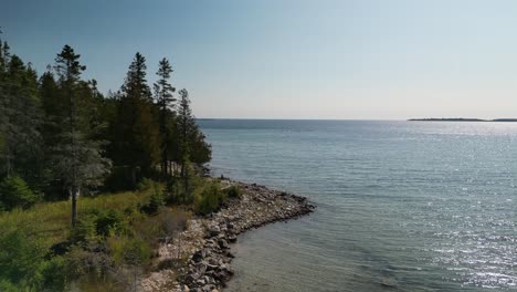 Aerial-view-up-shoreline-with-forest-and-shimmering-water,-Lake-Huron,-Michigan