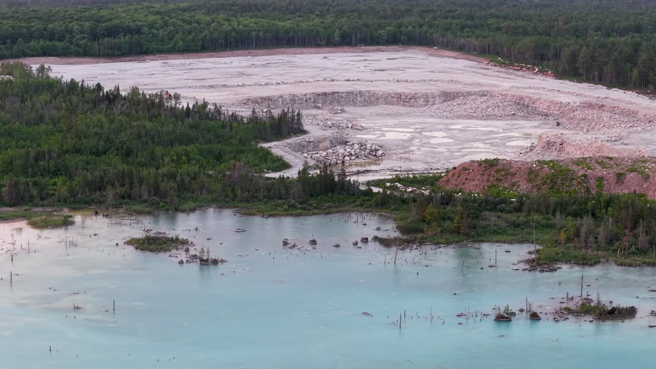 Aerial drone footage of a turquoise water quarry bordered by forest and a large exposed rocky mining area
