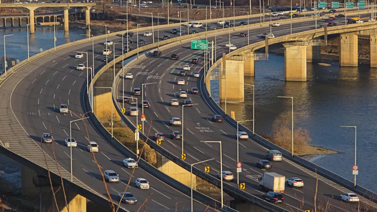 Detaillierte Echtzeit-Nahaufnahme von mehrspurigen, gekrümmten Gangbyeonbuk-ro-Autobahnrampen mit fließendem Verkehr über dem Han-Fluss, getaucht in warmes, goldenes Sonnenuntergangslicht.