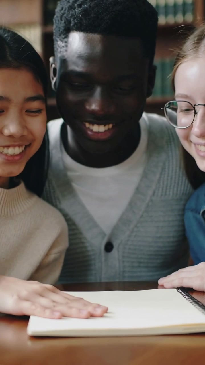Video verticale: Mano sinistra che si posa, studente a sinistra che traccia su un quaderno a spirale in biblioteca mentre gli amici studiano.