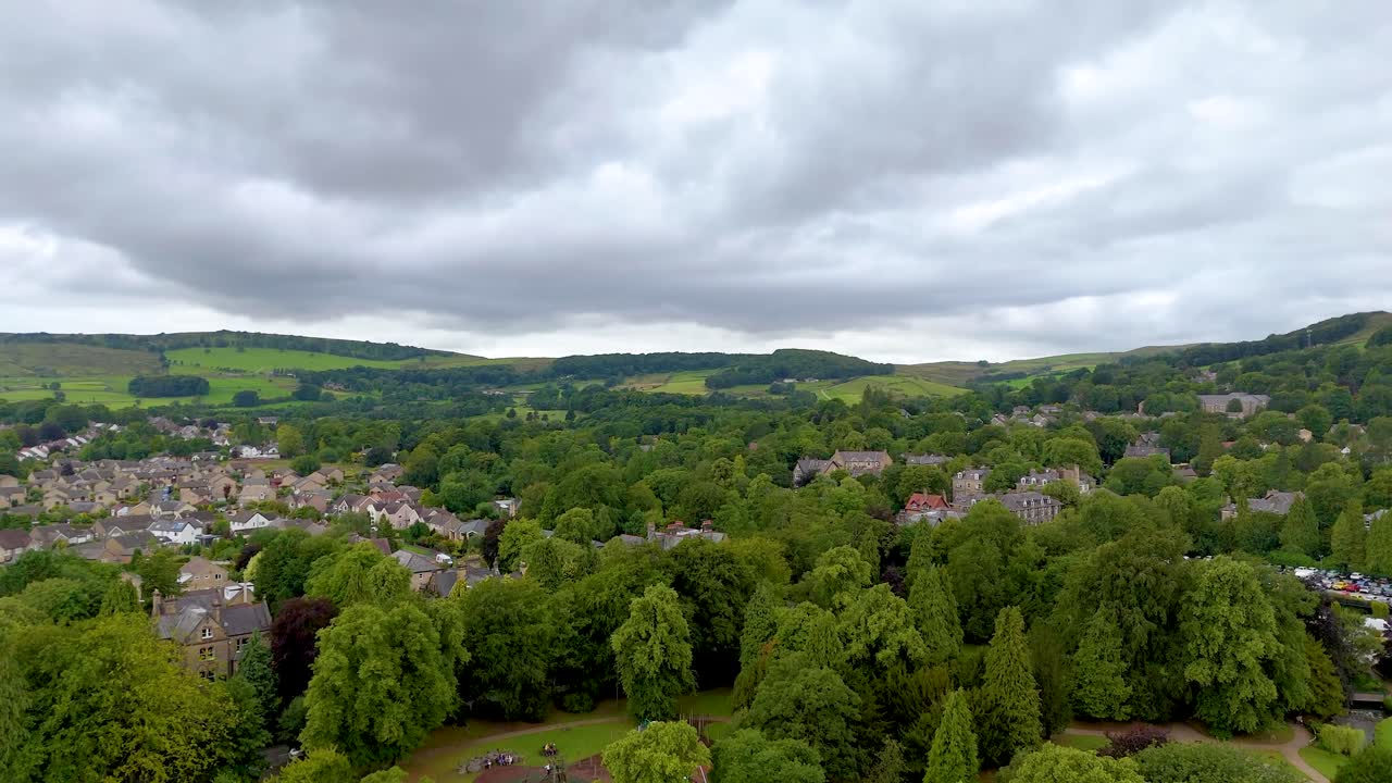 Aerial drone camera rises above lush green trees in a park, gradually revealing a small English town and rolling countryside under overcast skies