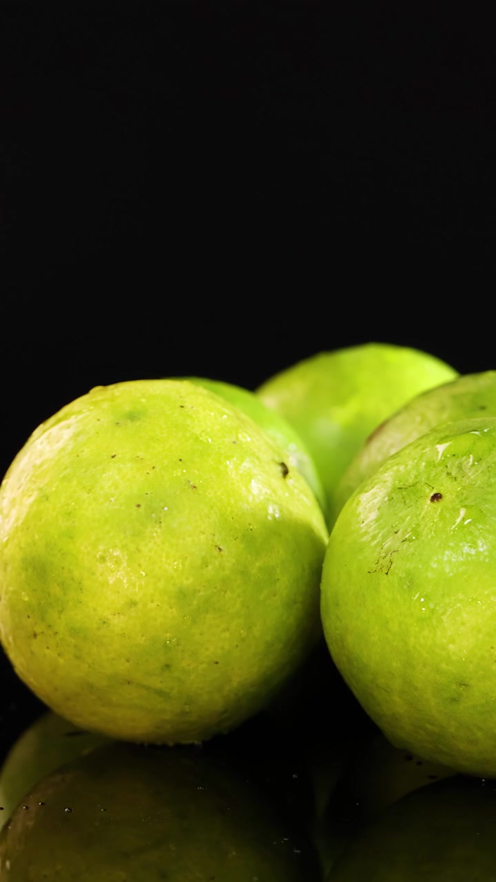 Um grupo de limas inteiras gira lentamente sobre uma superfície preta reflexiva, iluminado por uma luz de estúdio brilhante, realçando a sua textura fresca e cor verde vibrante.
