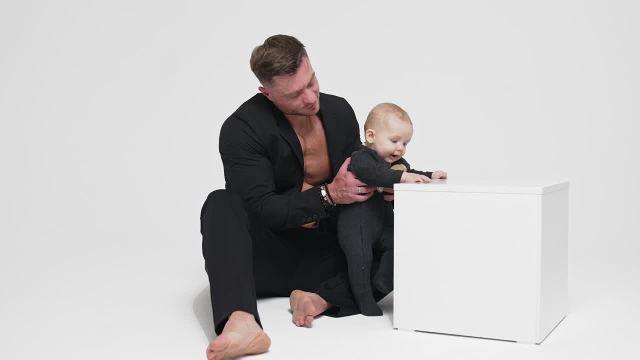 Strong muscular man in black jacket on bare chest holds a baby. Father and son studio footage. White backdrop.