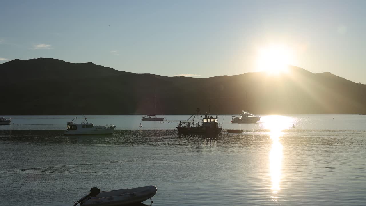 Calm waters with boats silhouetted against a rising sun in Akaroa, New Zealand. Warm lighting creates a serene atmosphere