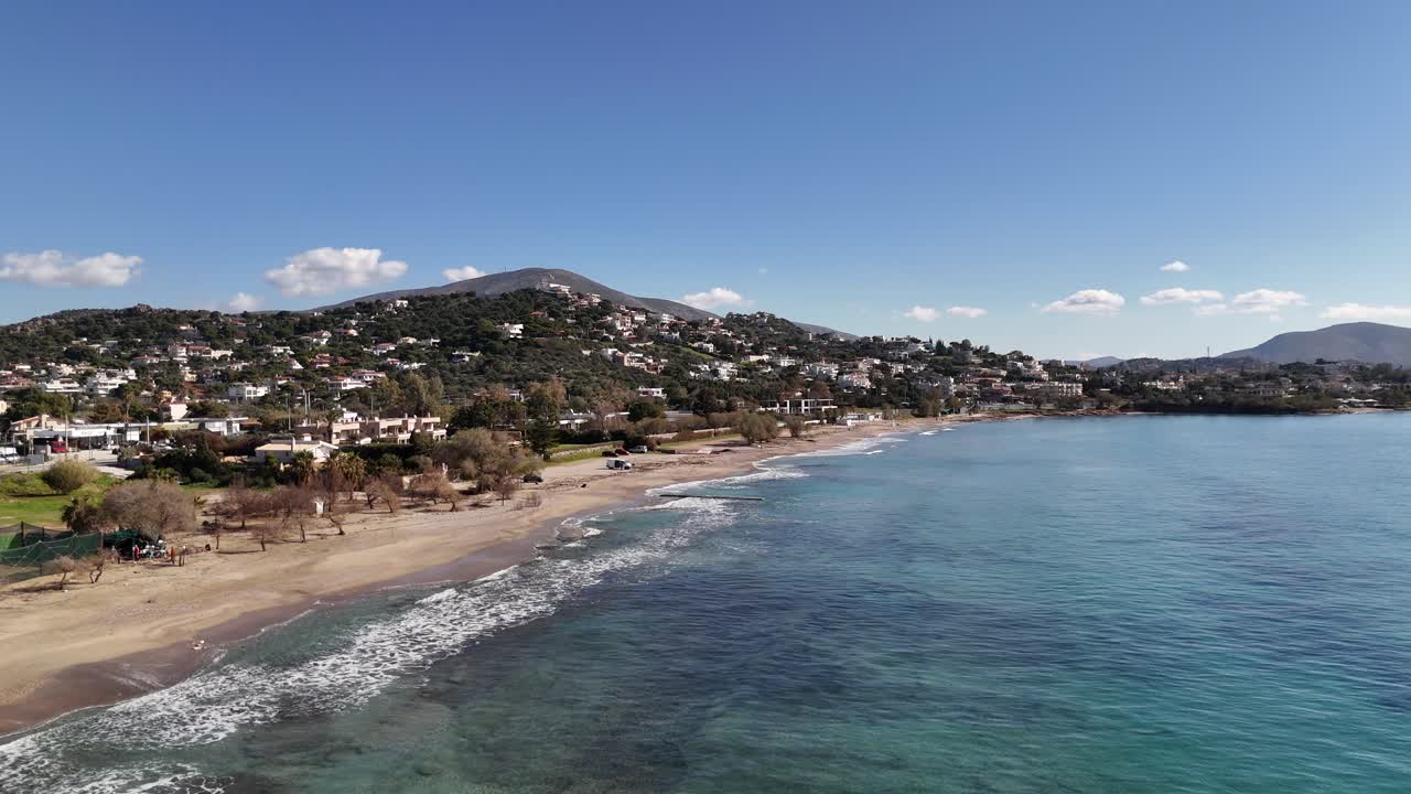 Vista aerea di una città costiera con mare turchese, spiaggia sabbiosa e case in collina sotto un cielo azzurro e limpido