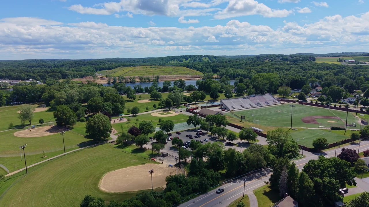 Campi da baseball e campi sportivi verdi riempiono il complesso del parco di Haverhill, parcheggio e alberi sparsi si estendono verso un piccolo stagno sotto le colline boscose del Massachusetts. Drone aereo in avvicinamento.