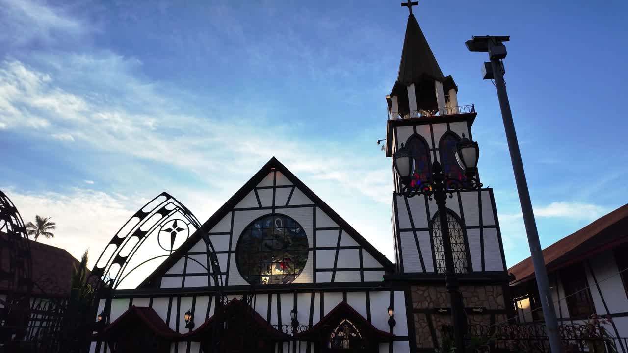 Fluida ripresa a carrello all'indietro che rivela una pittoresca chiesa in stile Tudor con intelaiature in legno sotto un cielo blu vibrante in un villaggio storico.
