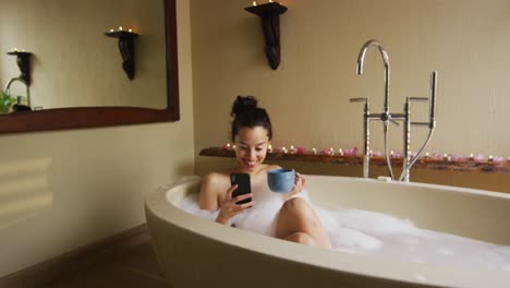 Happy-biracial-woman-taking-bath-with-foam,-drinking-coffee-and-using-smartphone