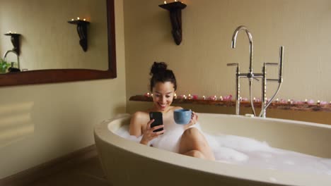 happy biracial woman taking bath with foam, drinking coffee and using smartphone