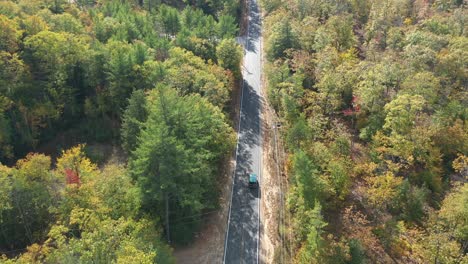 birdseye tracking aerial view of van moving on straight countryside road in fall landscape and colorful forest on sunny day, drone shot