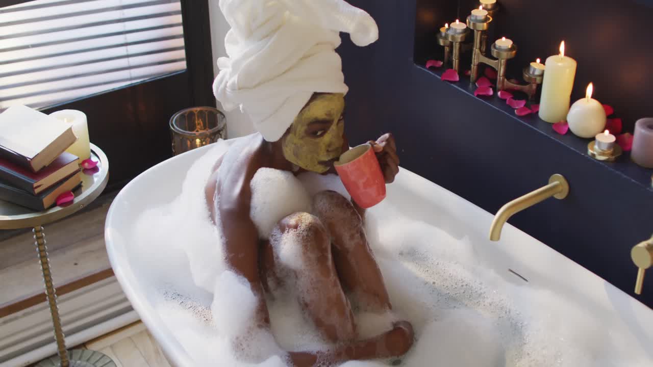 수건과 마스크를 착용 한 아프리카계 미국인 여성이 욕실에서 목욕하고 커피를 마시고 있습니다.