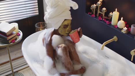 african american woman with towel and mask taking bath and drinking coffee in bathroom