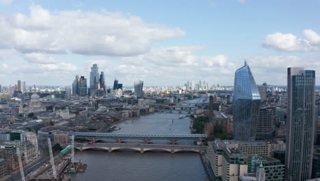 Forwards-fly-above-River-Thames.-Aerial-view-of-tall-modern-shiny-skyscrapers-on-both-banks.-Several-bridges-spanning-water-in-city.-London,-UK