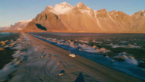 Aerial-view-orbiting-vehicle-on-long-road-leading-towards-Vestrahorn-Mountain-at-sunrise
