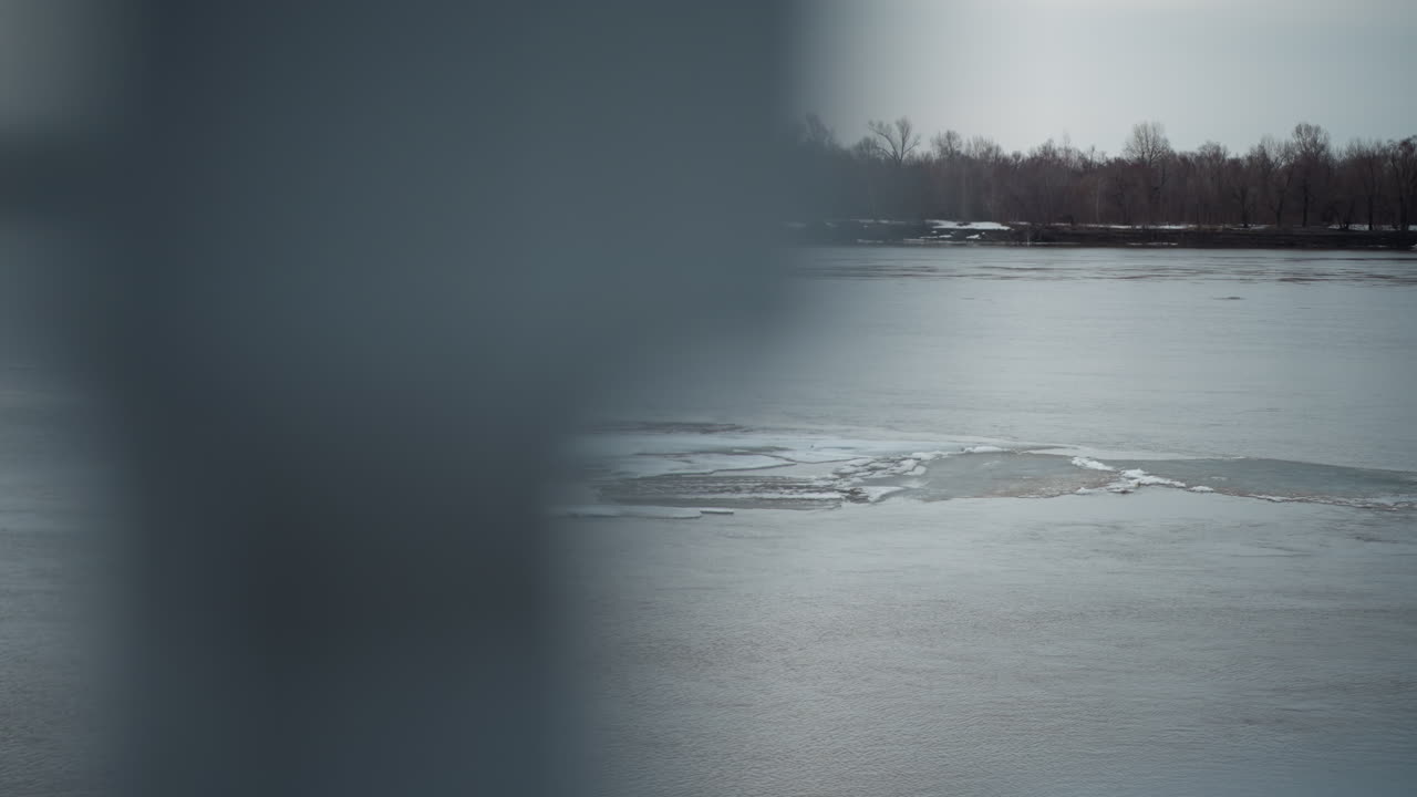 Frozen river with patches of ice floating on surface during cold winter day, distant shoreline lined with bare trees under overcast sky, small boat visible in background moving across tranquil water