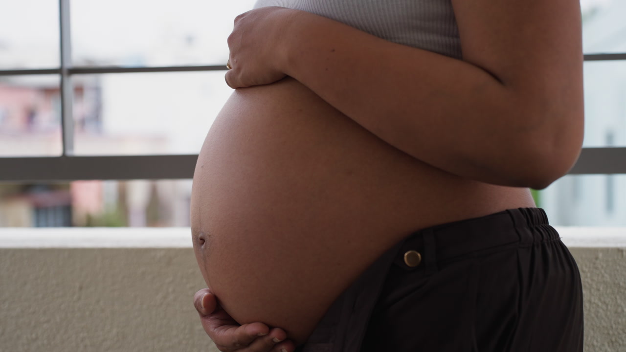 Pregnant Indian woman stands gently holding belly, serene outdoor moment in balcony terrace