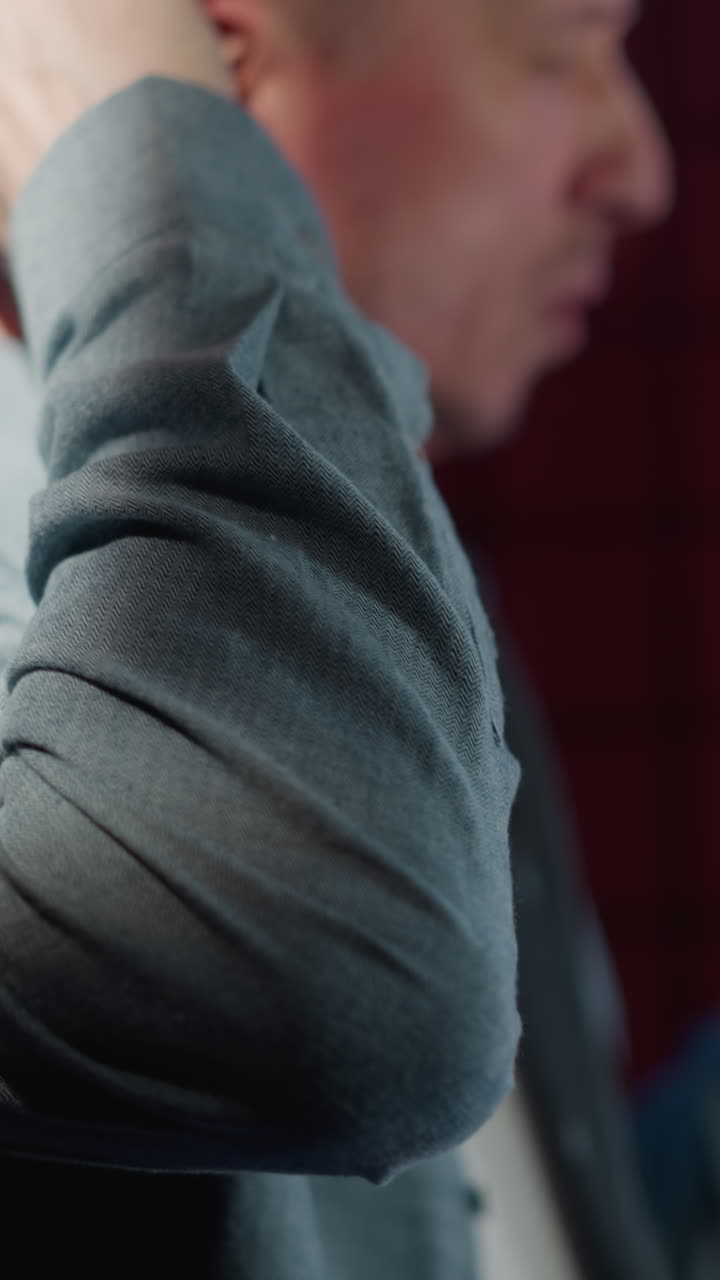 un primer plano de un hombre en un estudio de grabación con una camisa de manga larga azul, ajustando sus auriculares cantando en un micrófono, con un fondo de panel acústico rojo. el enfoque está en los movimientos de sus manos