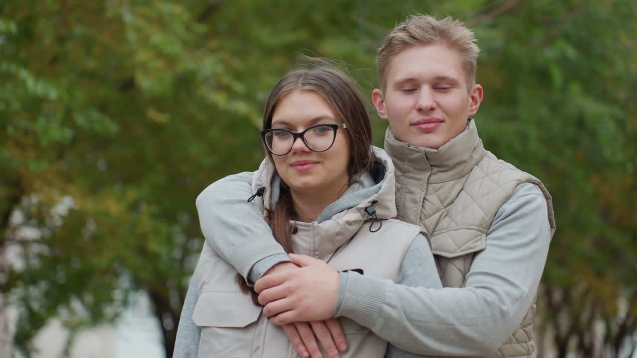 Young husband places hand across wife shoulder as they stand with eyes closed in peaceful moment, wearing matching outfits with tree branches swaying gently in wind
