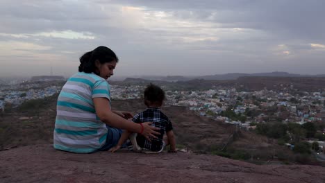 Isolierte-Mutter-Und-Kleiner-Sohn-Beobachten-Die-Stadtlandschaft-Auf-Dem-Berggipfel-Mit-Dramatischem-Himmel-In-Der-Abenddämmerung.-Das-Video-Wurde-In-Mehrangarh,-Jodhpur,-Rajasthan,-Indien,-Aufgenommen