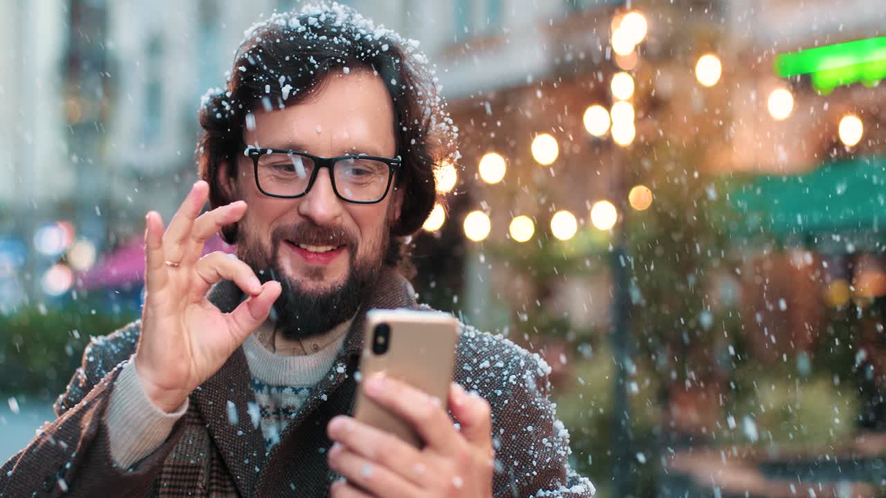 Close-up view of caucasian man wearing eyeglasses making a video call with smartphone on the street while it‚Äôs snowing in Christmas