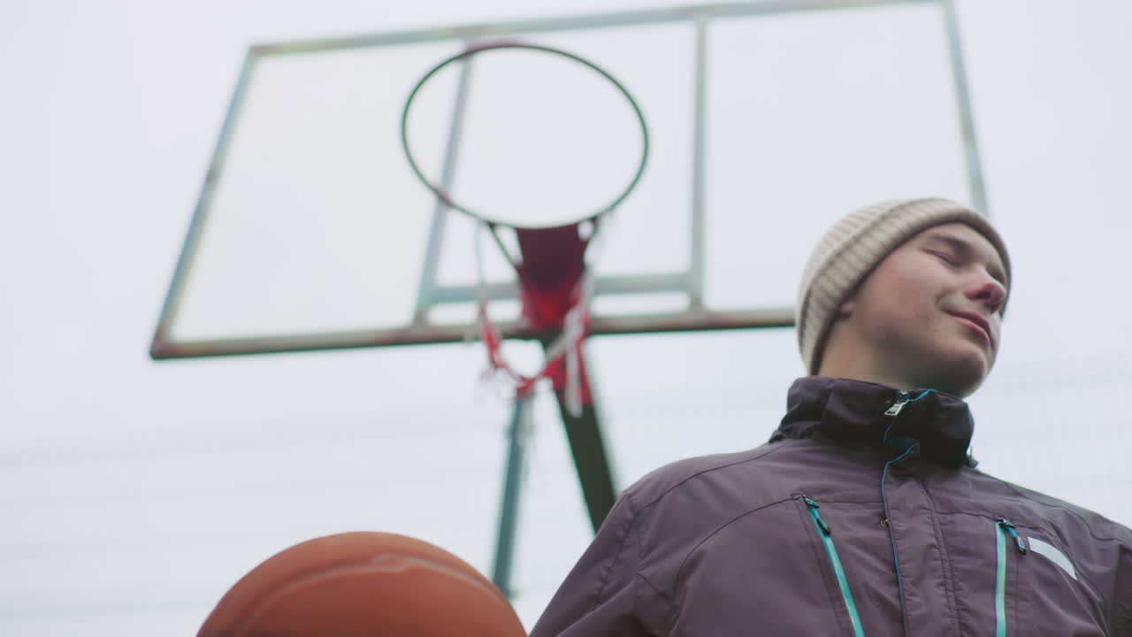 Ritratto dal basso che mostra un ragazzo bianco adolescente sotto un canestro da basket, con un pallone in mano, che indossa un berretto e una giacca invernale su un campo urbano nuvoloso; atleta di strada solitario e sicuro di sé che si prepara a giocare