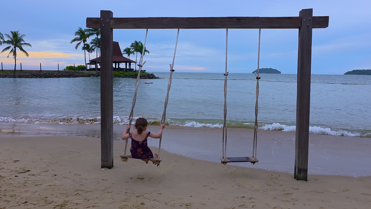 Little happy girl swinging on a tropical beach at sunset. Cute blonde kid girl is swinging on a sandy beach by the ocean in the tropical island of Kota Kinabalu