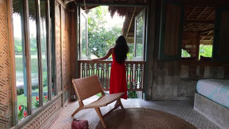 asian woman with long red dress looking out the balcony window in a traditional bohemian villa in bali