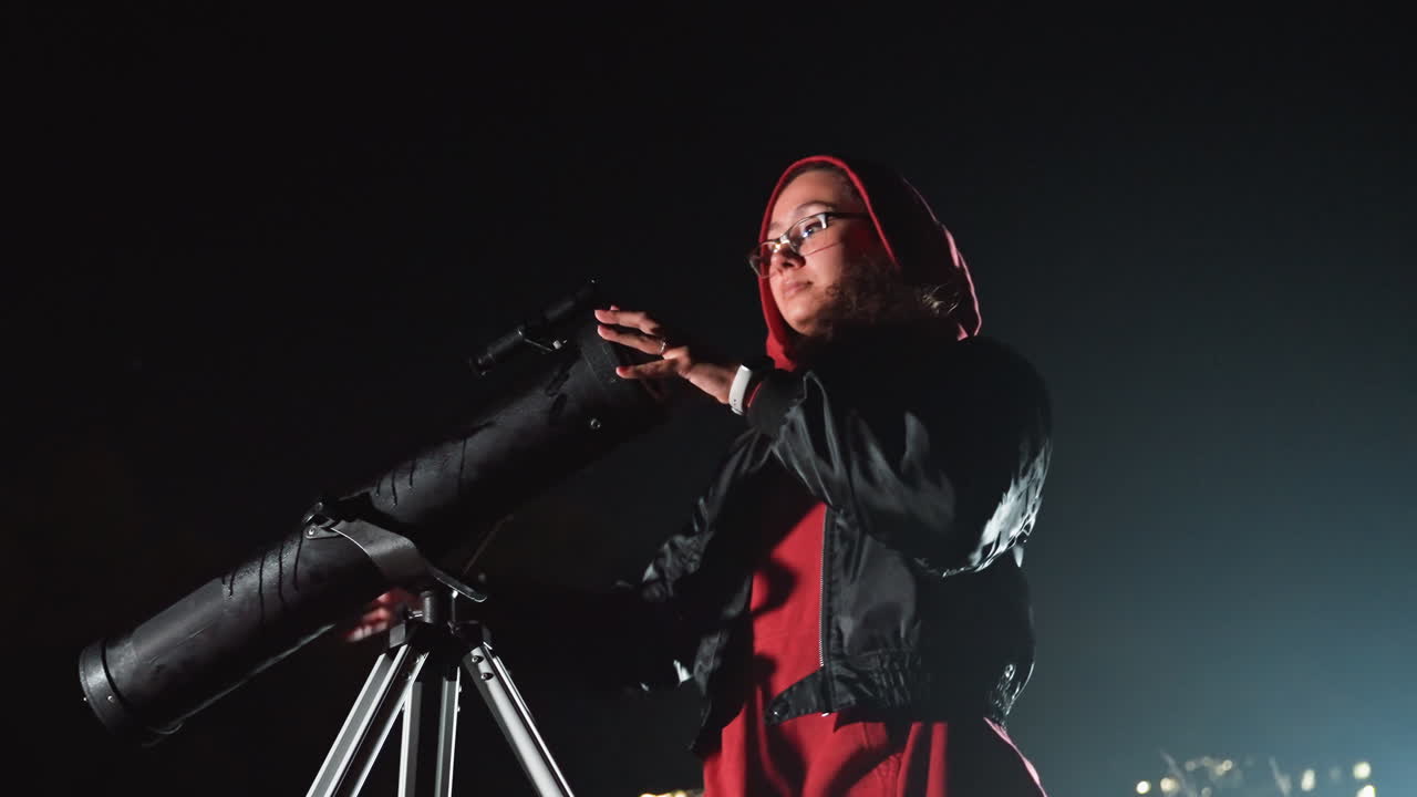 Donna su un tetto di notte con un telescopio che regola l'oculare sotto le luci della città, giacca di pelle e cappuccio, sguardo pensieroso verso il cielo, telescopio montato su un treppiede puntato alle stelle, mani che mostrano la tecnica