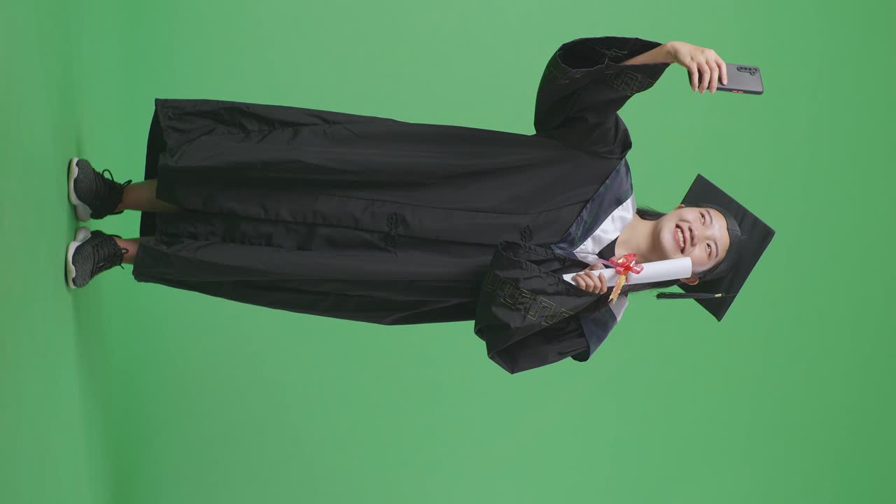 Studentessa asiatica laureata a figura intera con tocco e toga e diploma, che scatta una foto con lo smartphone mentre è in piedi su uno sfondo green screen in studio.