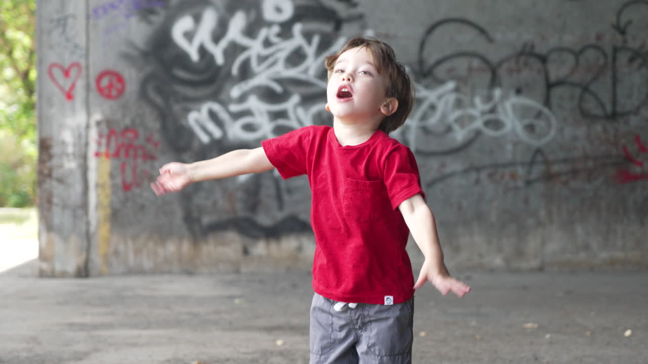 Slow motion video of a young little boy spot jumping in front of camera in red T-shirt, Young boy playing in front of a graffiti wall in a abandoned place