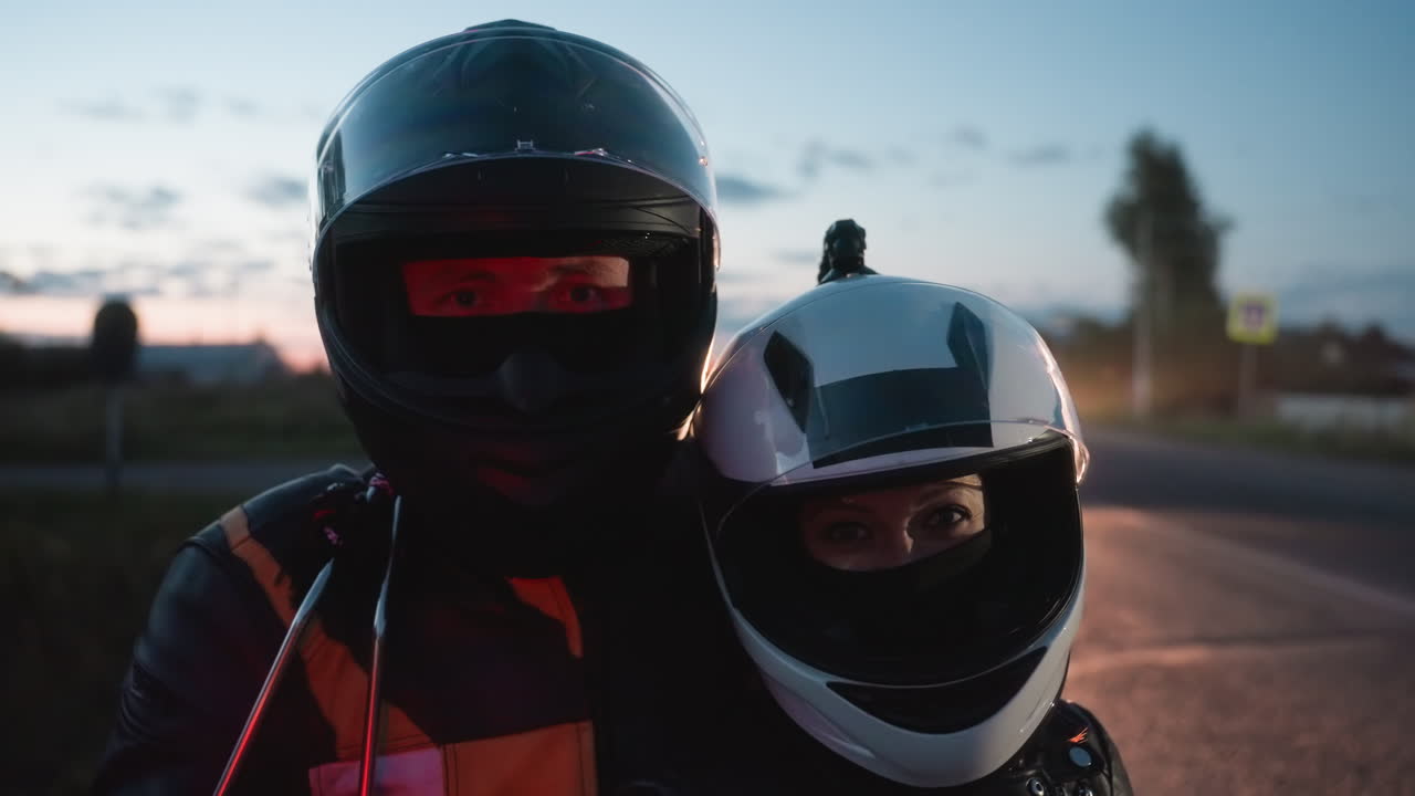 Close up of man and woman in helmets and leather jackets standing together near motorcycle at roadside during sunset as passing car headlights illuminate them