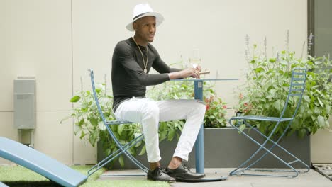 hombre rico con sombrero fedora y joyas de oro se sienta en una mesa bebiendo vino afuera cerca de plantas verdes